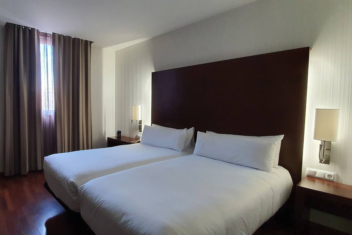 Ein Hotelzimmer mit zwei Betten, einem Schreibtisch, einem Stuhl, einer Lampe und einem Fenster mit Vorhängen.