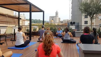 Un grupo de personas sentadas sobre colchonetas de yoga en una terraza de madera, con vistas a un paisaje urbano de fondo.