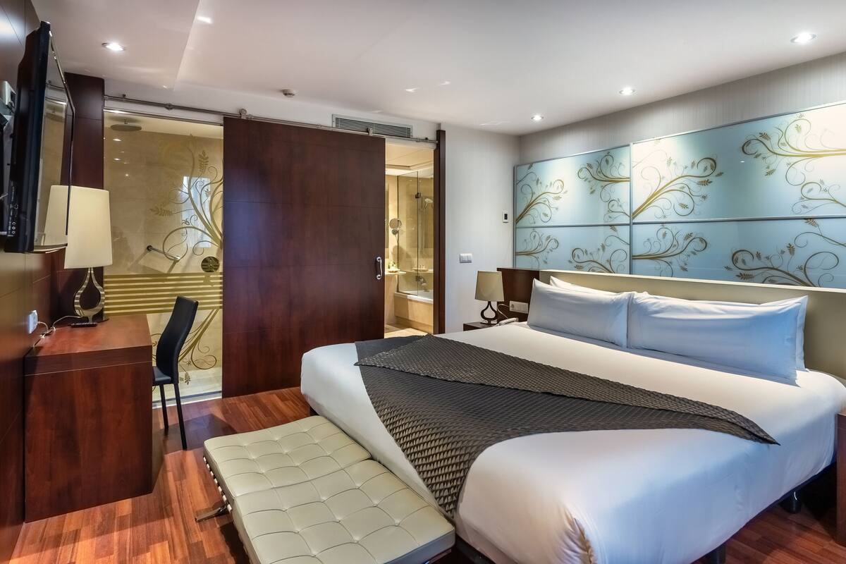 Ein modernes Hotelzimmer mit einem großen Bett, einem Schreibtisch mit Lampe und einem durch eine Schiebetür einsehbaren Badezimmer.