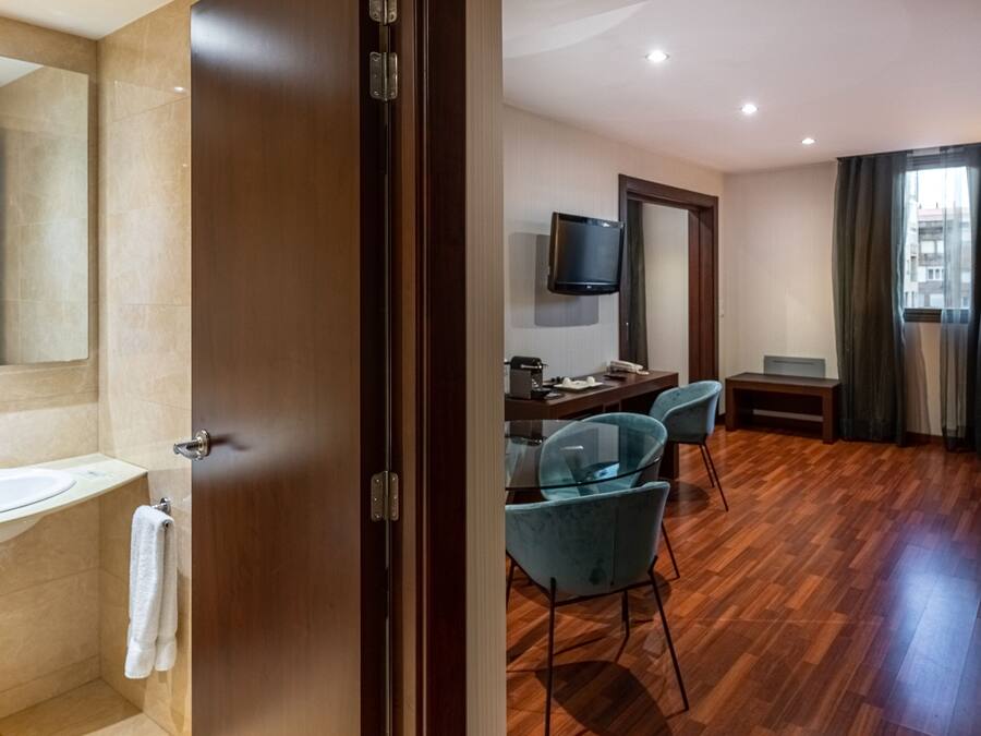 Habitación de hotel con piso de madera, una cama, un escritorio con silla, televisor montado en la pared y un ventanal con cortinas.