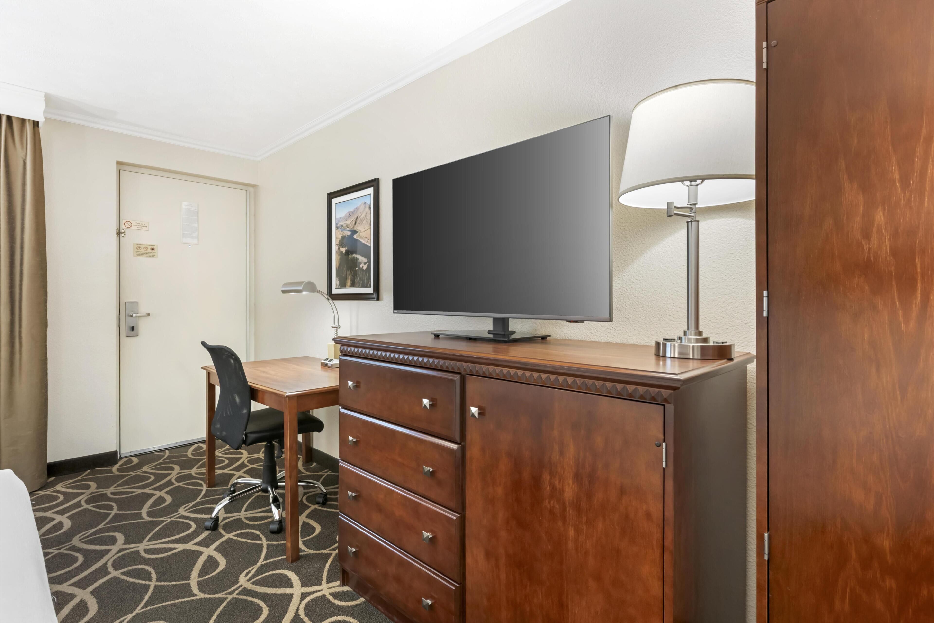 A hotel room with a desk, chair, television, dresser, and a lamp.