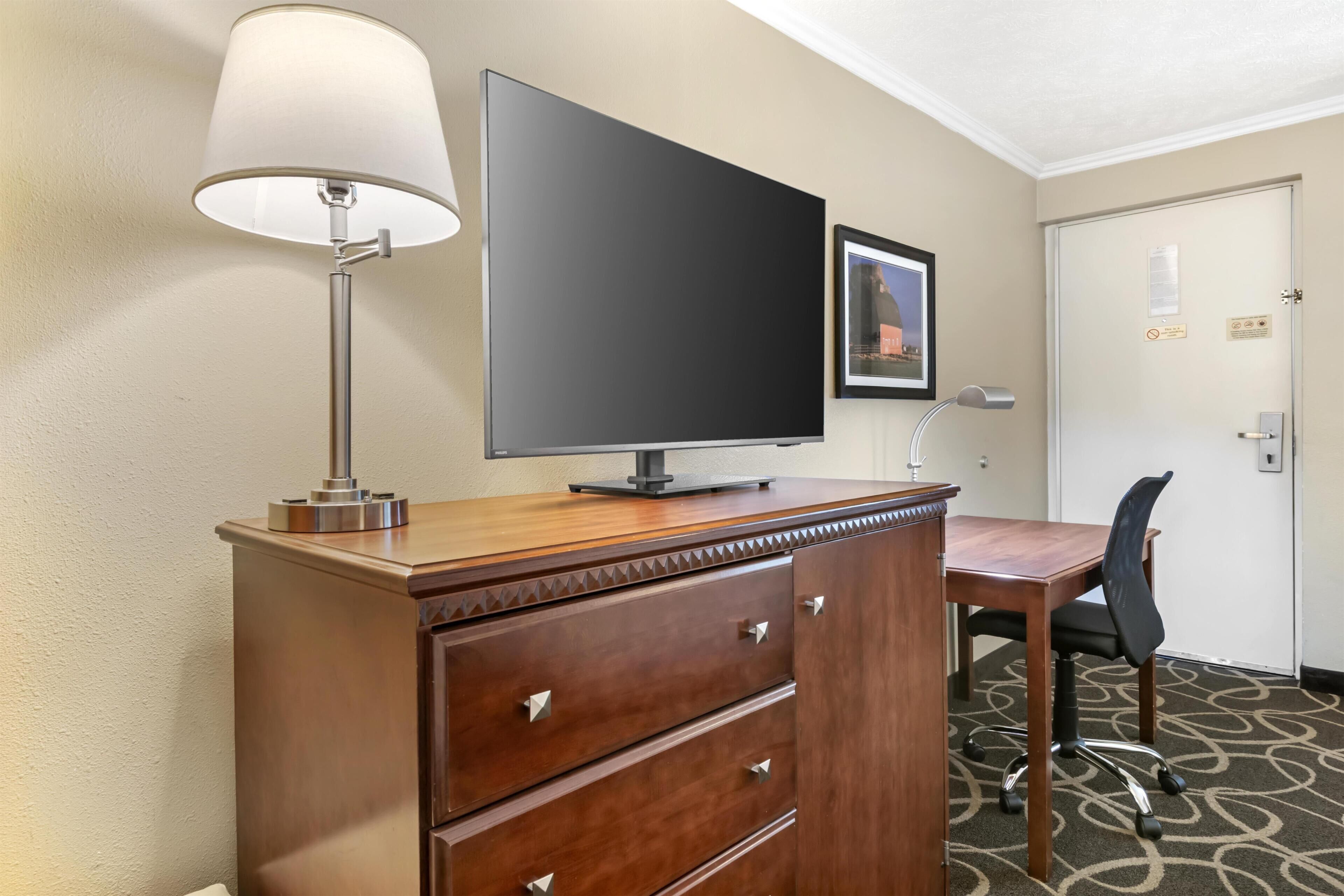 A hotel room with a wooden desk, a flat-screen TV, a lamp, a chair, and a door with a picture frame and a sign.