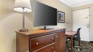 A hotel room with a wooden desk, a flat-screen TV, a lamp, a chair, and a door with a picture frame and a sign.