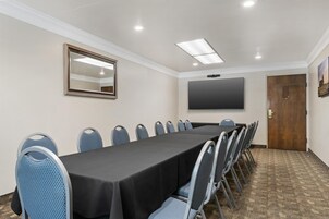 Uma sala de conferências com uma mesa comprida, cadeiras, um espelho grande, uma TV de tela plana e uma porta.