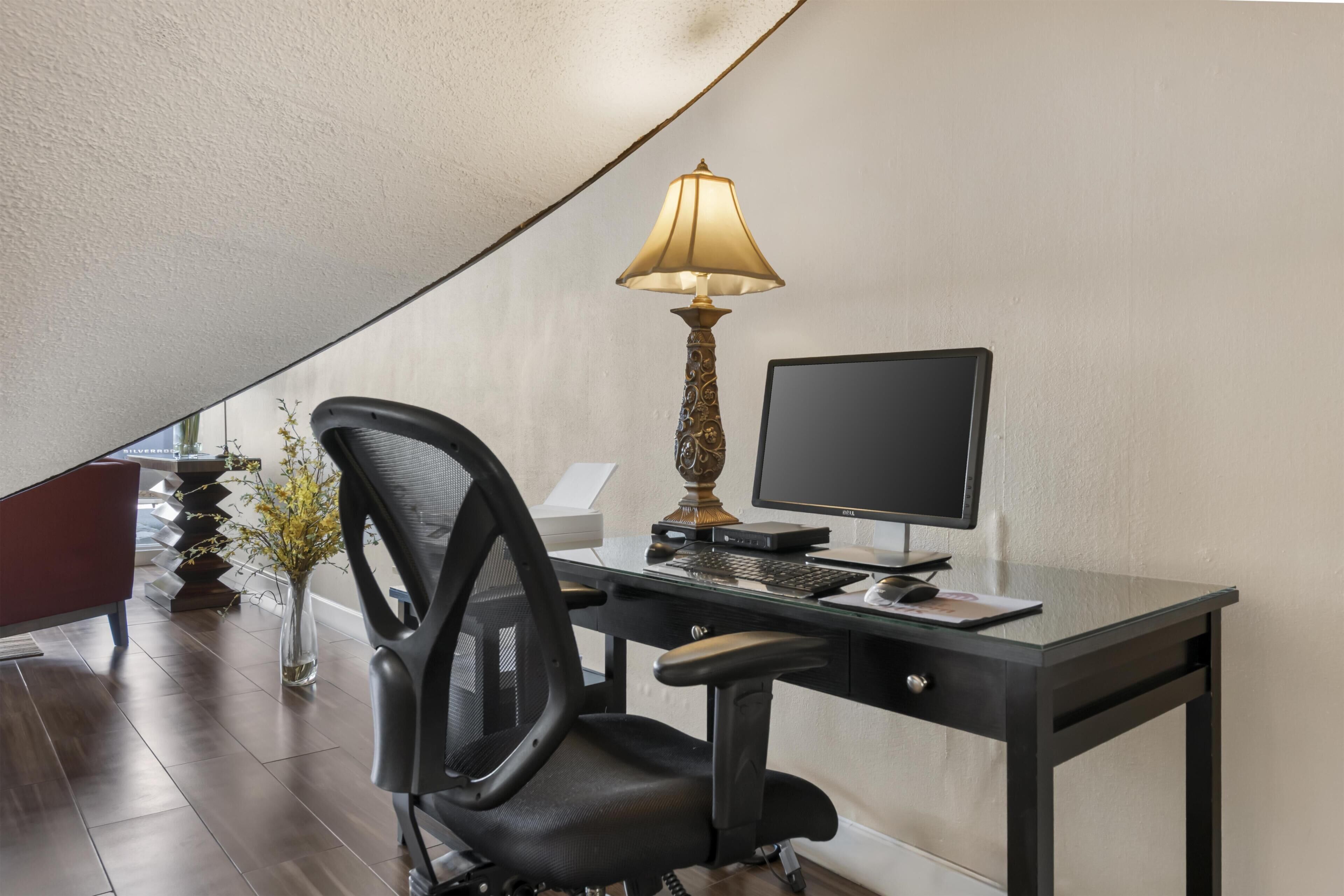 Un espacio de oficina moderno con escritorio, silla, computadora, lámpara y un florero con flores.