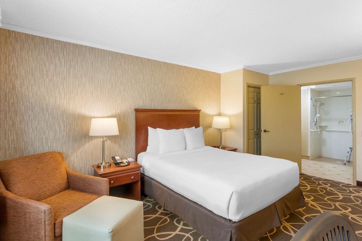 A hotel room with a bed, a chair, a nightstand, a lamp, and a door leading to a bathroom.