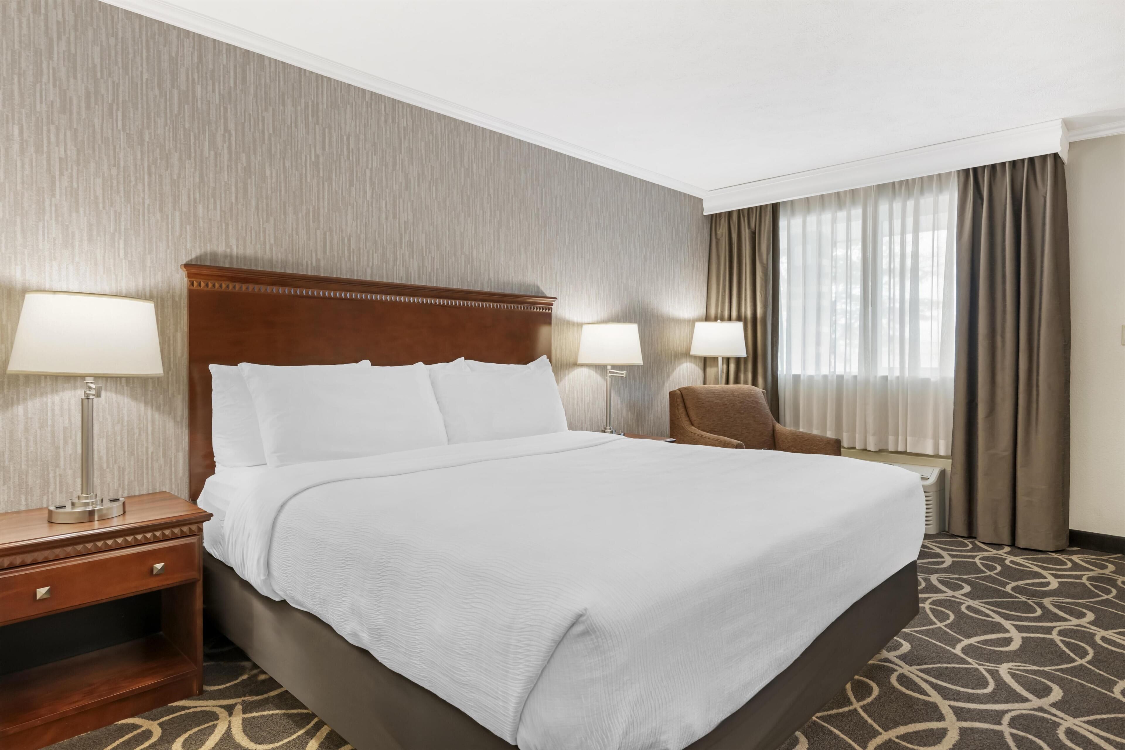 Una habitación de hotel con una cama grande, dos lámparas de noche, una silla y un ventanal con cortinas.