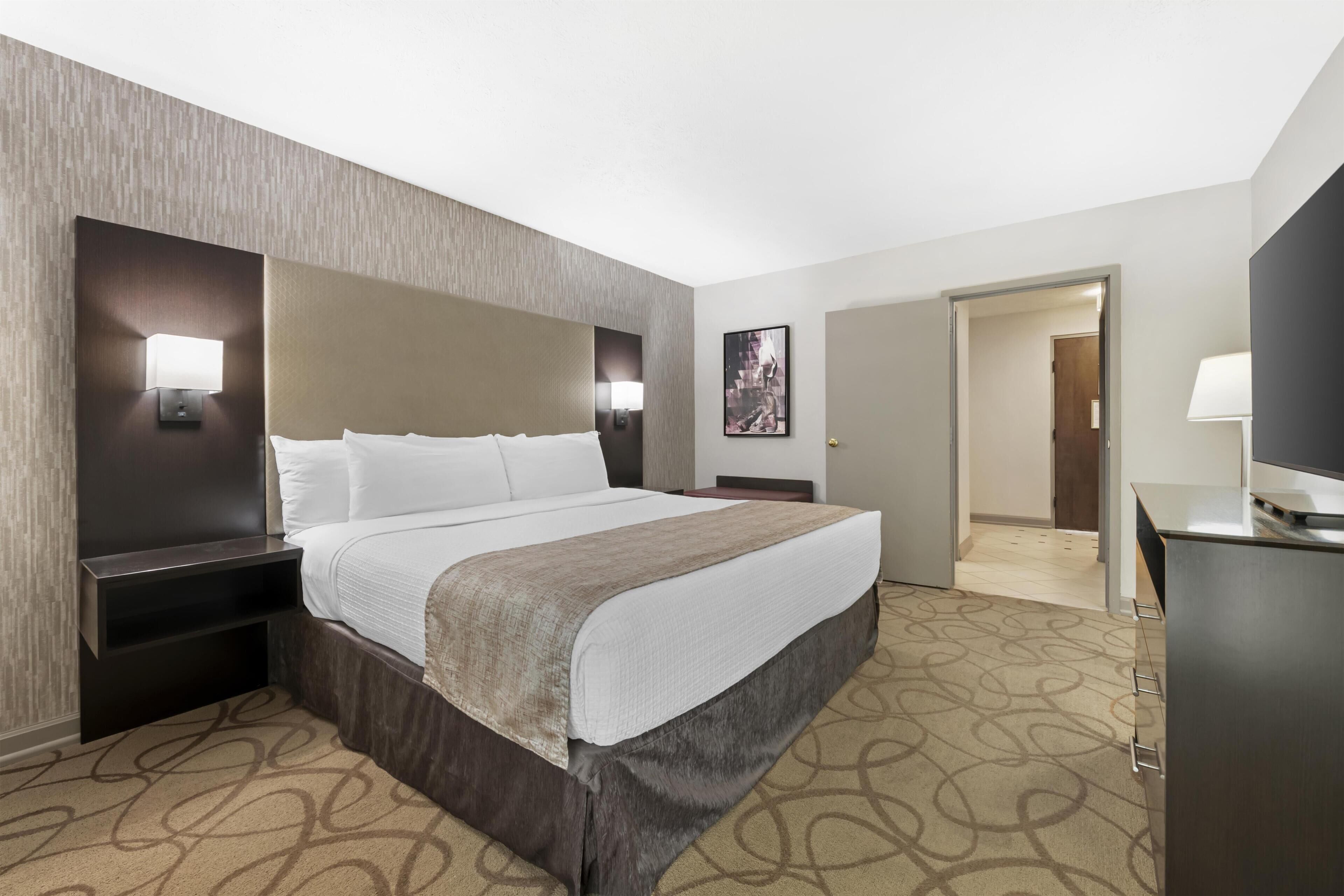 A hotel room with a large bed, bedside tables, a flat-screen TV, and a door leading to another room.