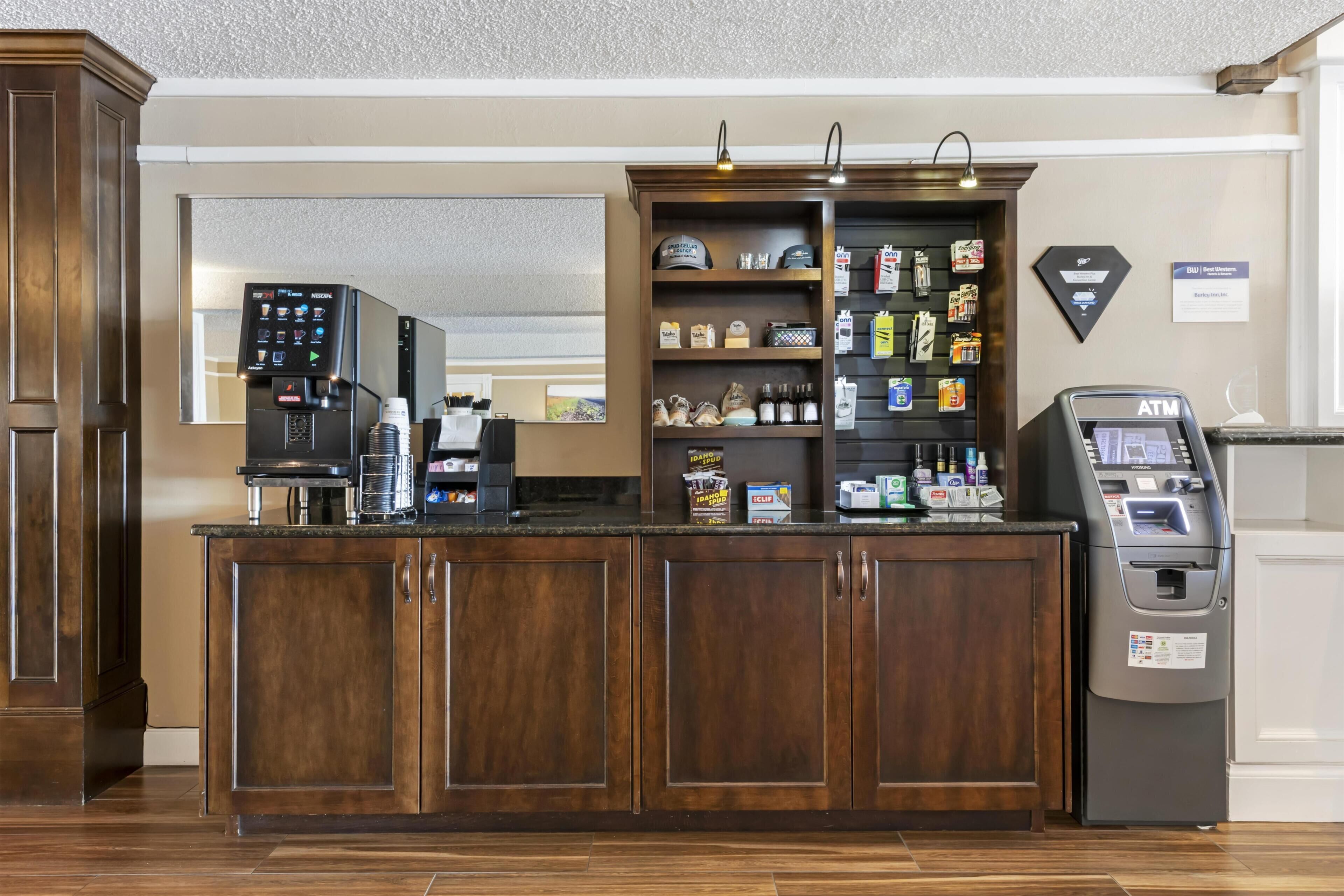 A hotel reception area with a coffee machine, a self-service kiosk, and a variety of snacks and toiletries on shelves.