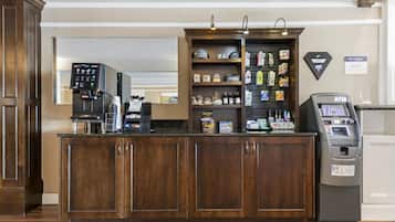 A hotel reception area with a coffee machine, a self-service kiosk, and a variety of snacks and toiletries on shelves.