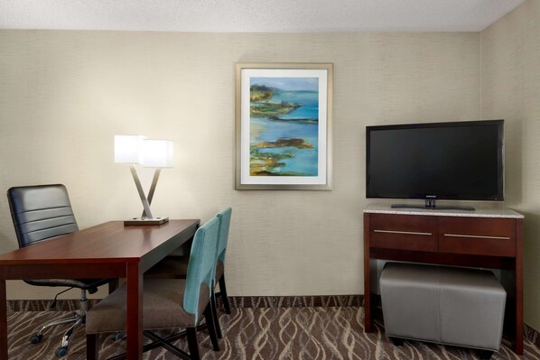 A hotel room with a wooden desk, a television, a painting on the wall, and a patterned carpet.