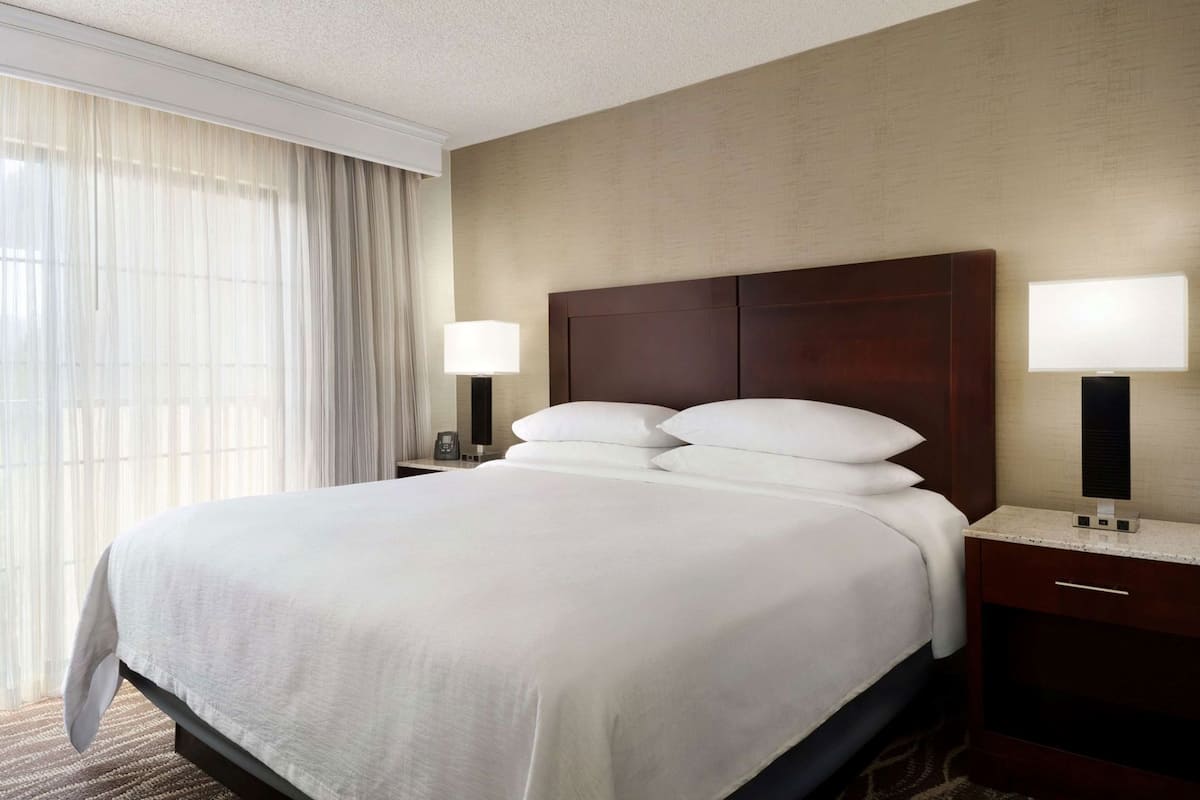 A hotel room with a large bed, two bedside lamps, a nightstand, and a window with sheer curtains.