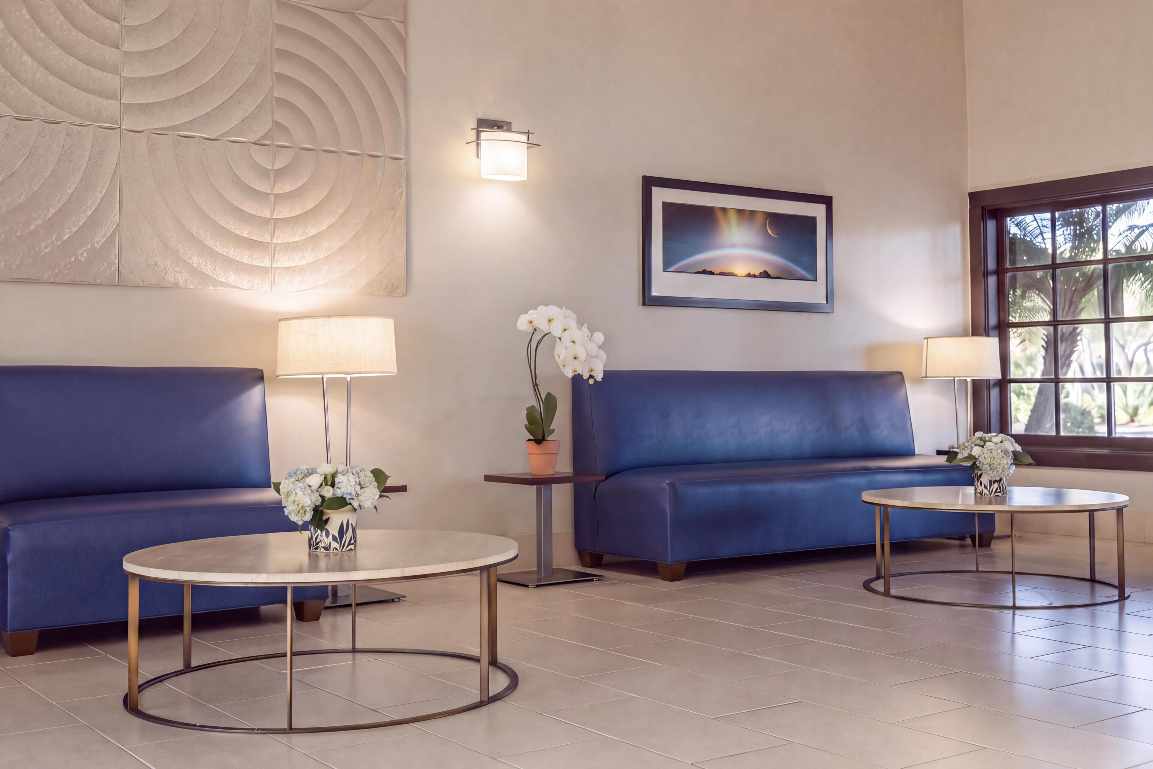 Un salon moderne avec un canapé bleu, deux tables rondes, une lampe fixée au mur, un tableau encadré et une fenêtre donnant sur des palmiers.