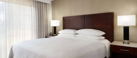 A hotel room with a large bed, two bedside tables with lamps, a wooden headboard, and a large window with sheer curtains.