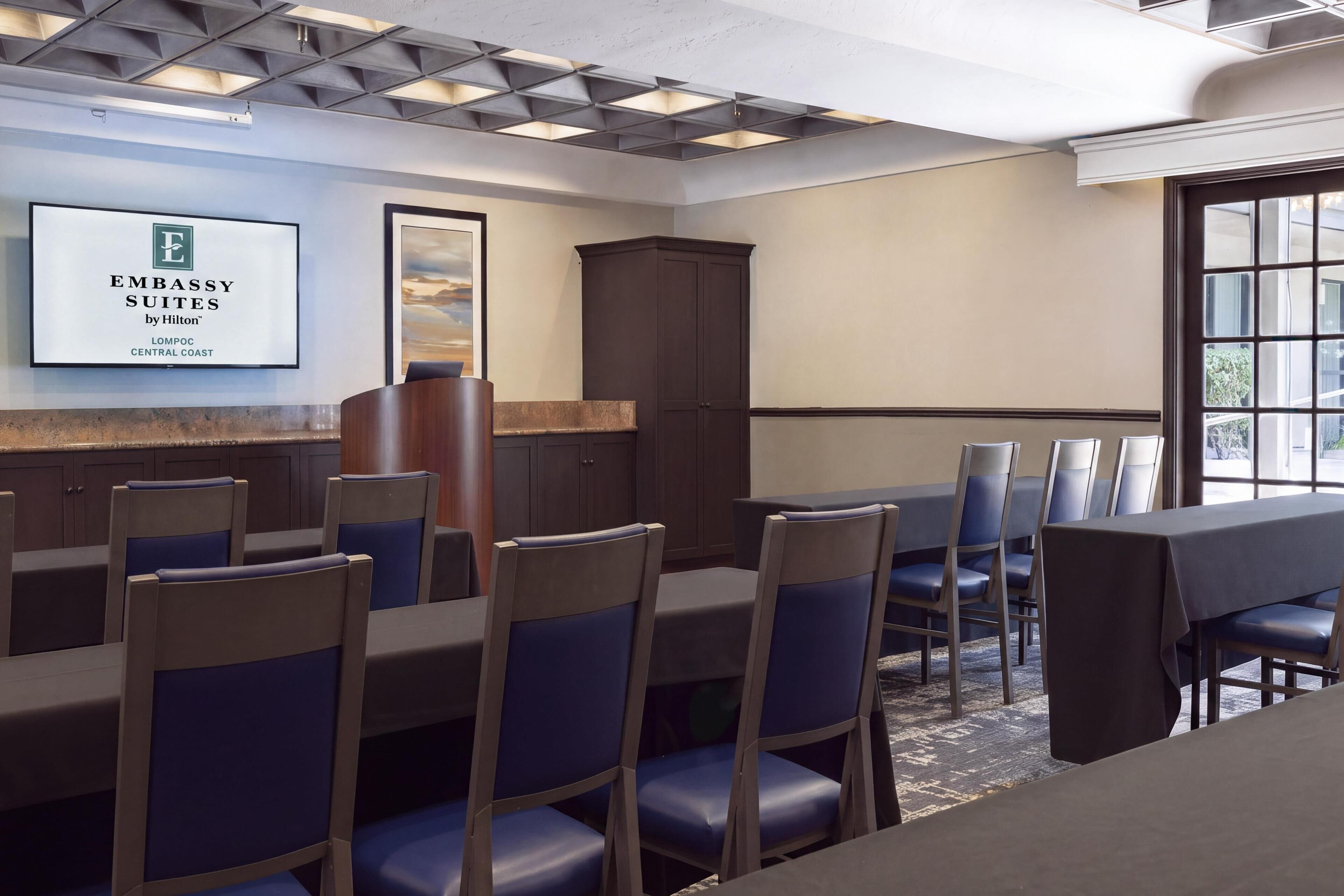 Uma sala de conferências de hotel com uma mesa grande, cadeiras, um armário e uma placa emoldurada na parede.