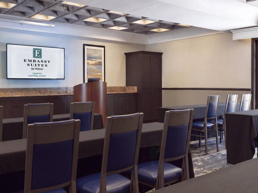 Uma sala de conferências de hotel com uma mesa grande, cadeiras, um armário e uma placa emoldurada na parede.