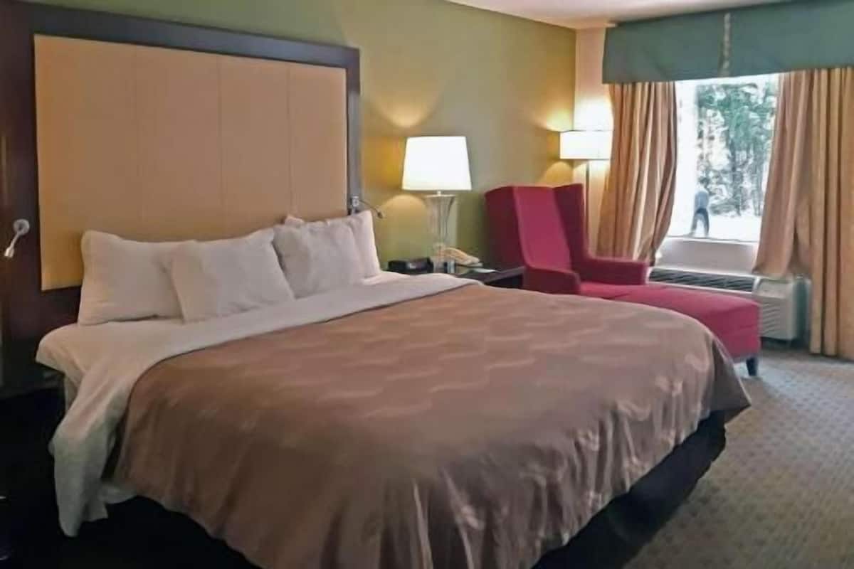 Habitació d'hotel amb un llit gran, una tauleta de nit, un escriptori, una cadira, una làmpada i una finestra amb cortines.