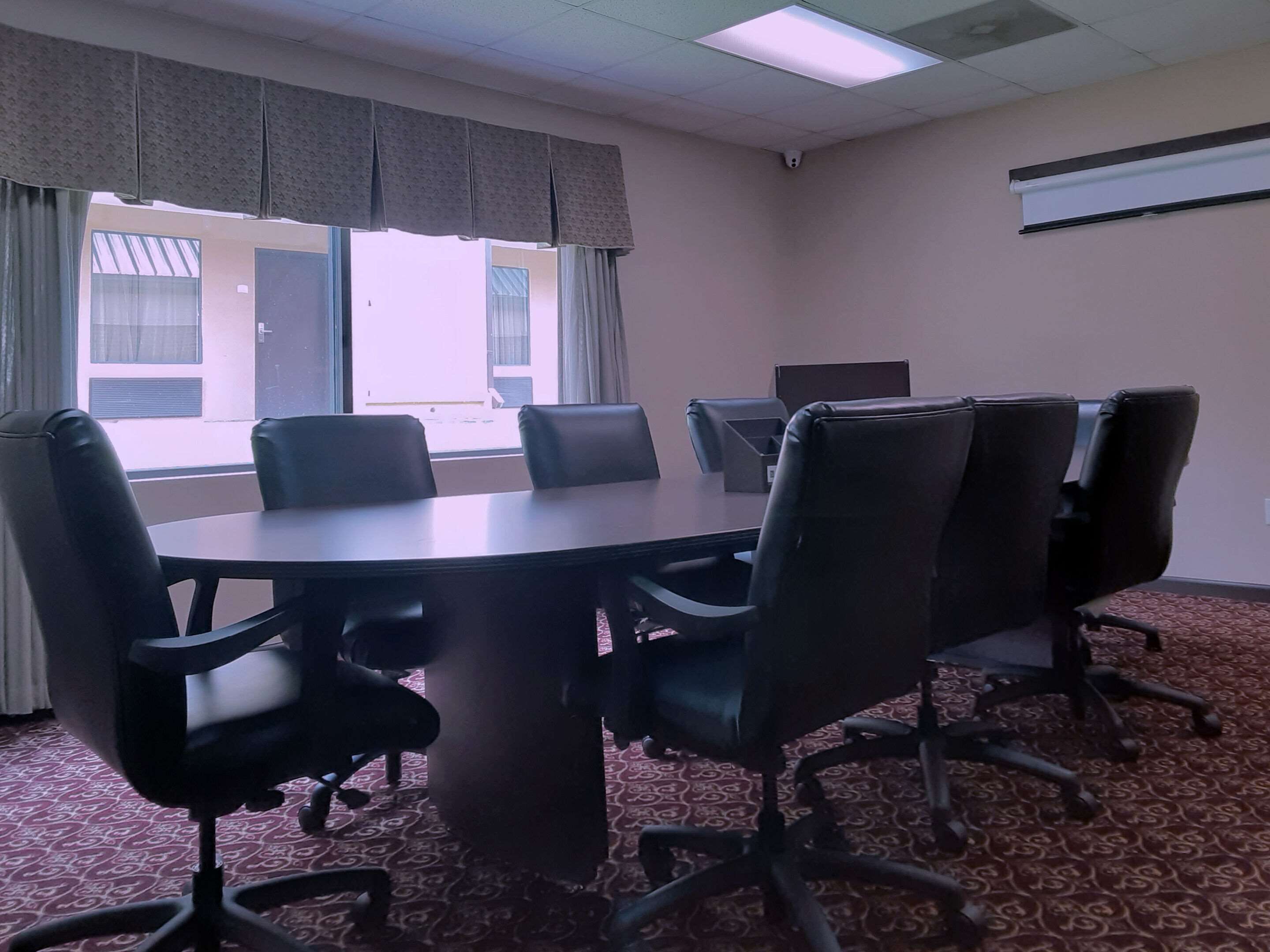 A conference room with a round table, black chairs, patterned carpet, large windows with curtains, and an air conditioning unit on the wall.