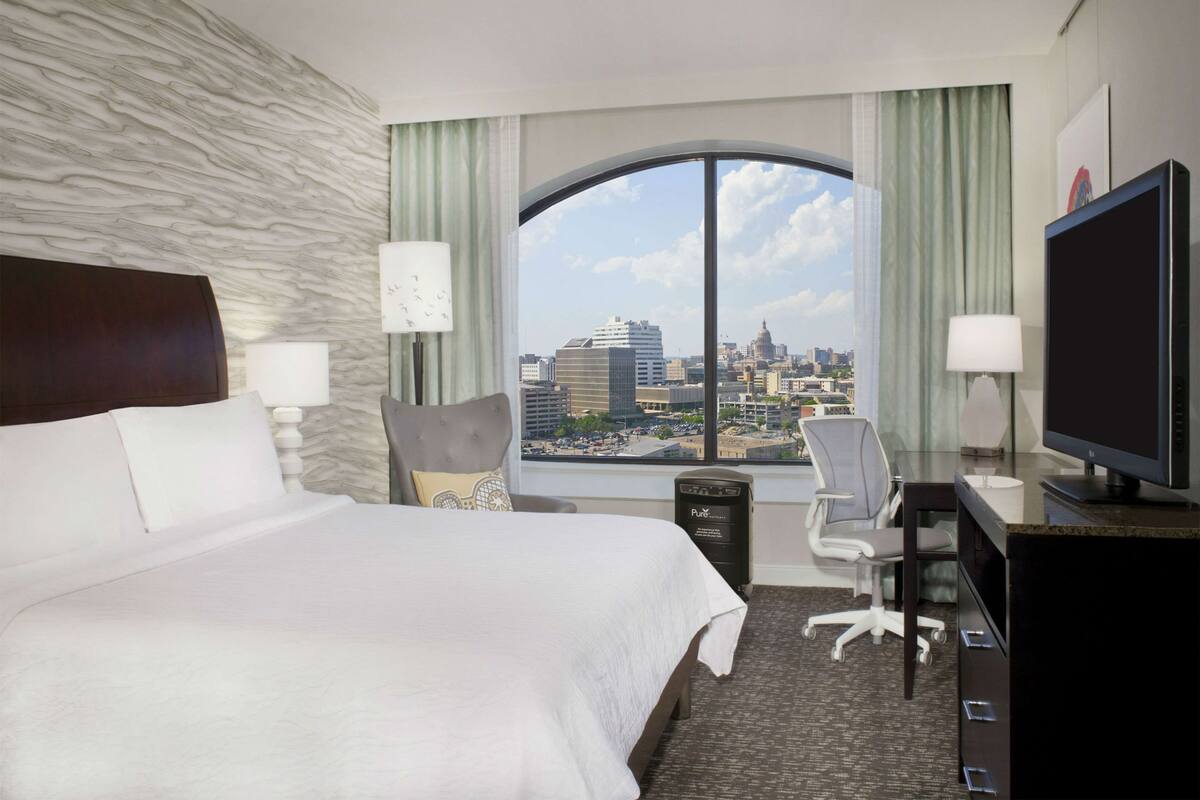 A hotel room with a large bed, a desk with a TV, a chair, and a view of a cityscape through a large window.