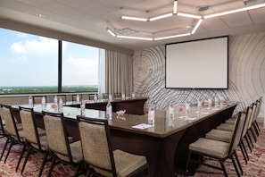 A conference room with a large table, chairs, a projector screen, and a view of the city. - Hilton Garden Inn Austin Downtown (Austin)