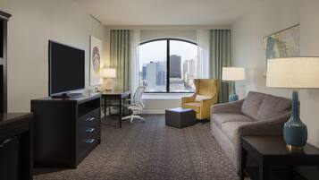 A hotel room with a desk, chair, sofa, and a large window offering a city view.