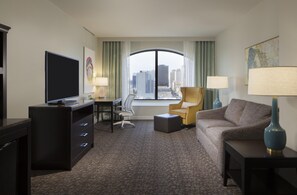 A hotel room with a desk, chair, sofa, and a large window offering a city view. - Hilton Garden Inn Austin Downtown (Austin)