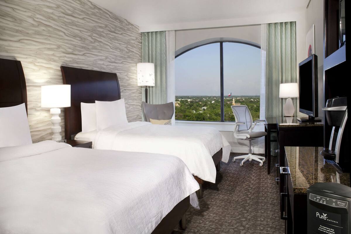 A hotel room with two beds, a desk, a chair, a TV, and a large window offering a city view.