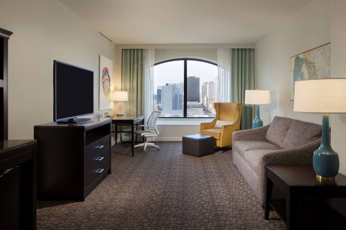 A hotel room with a large window, a desk with a computer, a sofa, a chair, and a TV.
