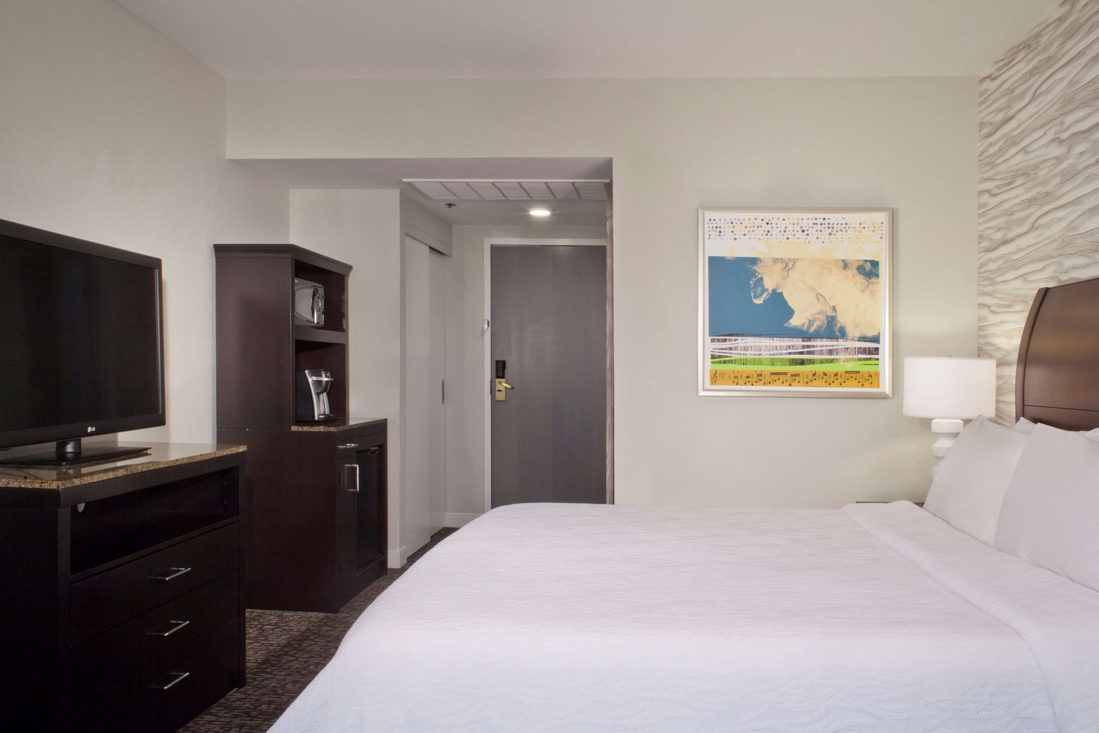 A hotel room with a bed, a television, a minibar, and a door leading to a hallway.
