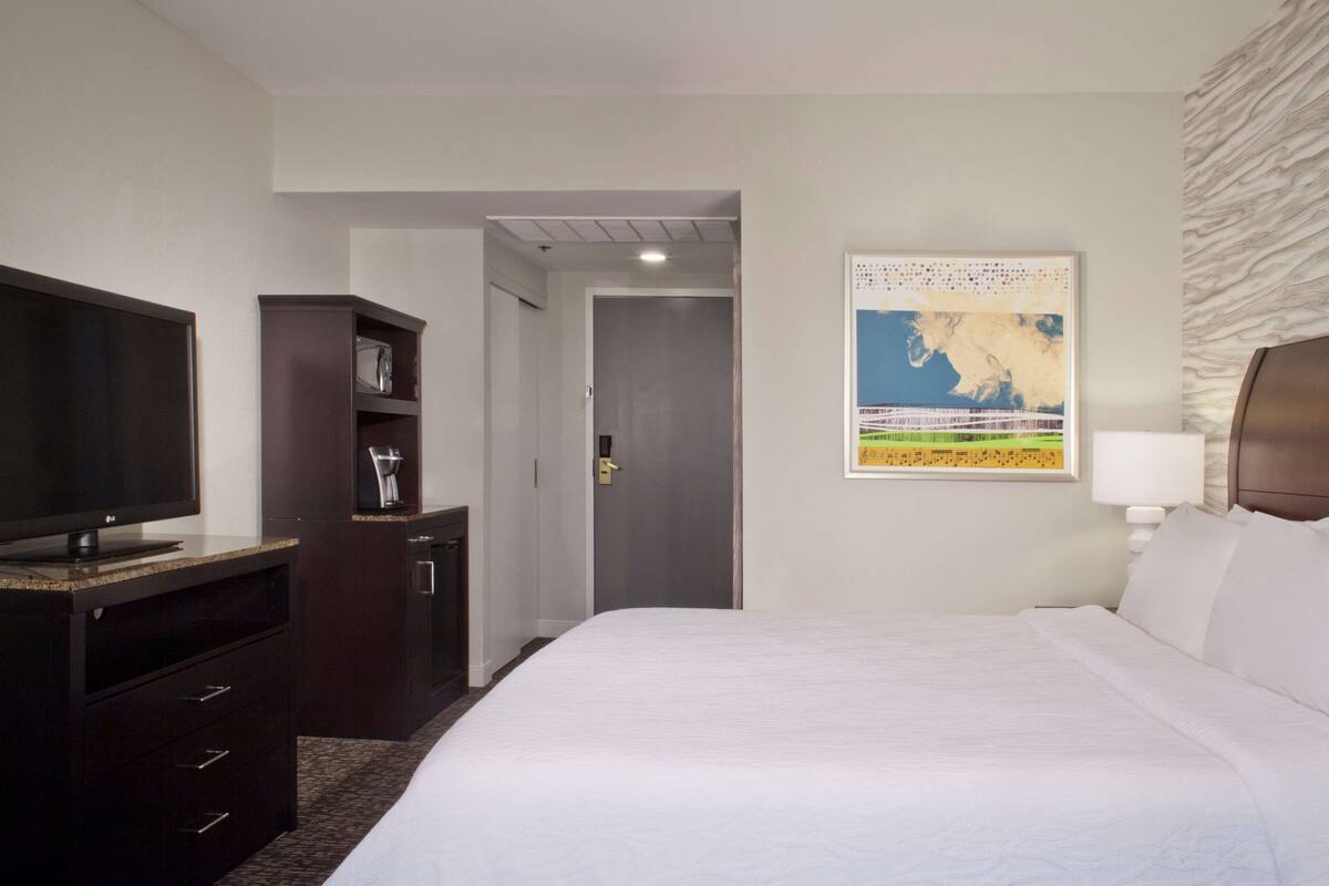 A hotel room with a bed, a television, a minibar, and a door leading to a hallway.