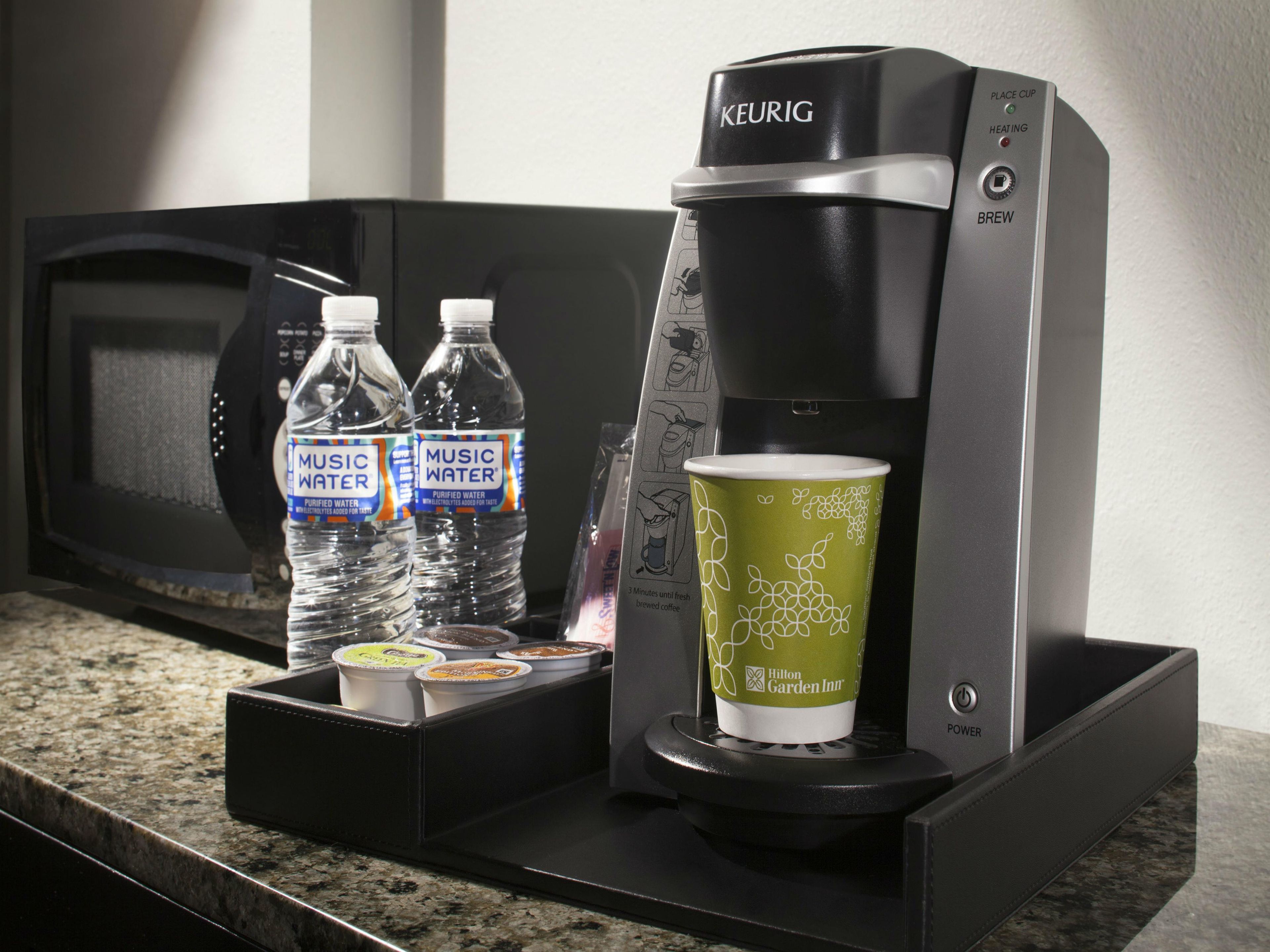 A Keurig coffee maker with a green cup, two bottles of Music Water, and a tray of coffee pods on a countertop.