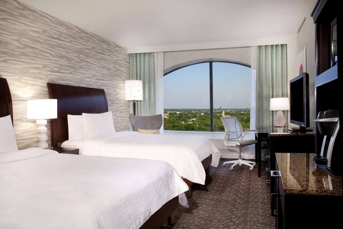 A hotel room with two beds, a desk, a chair, a TV, and a large window with a view.