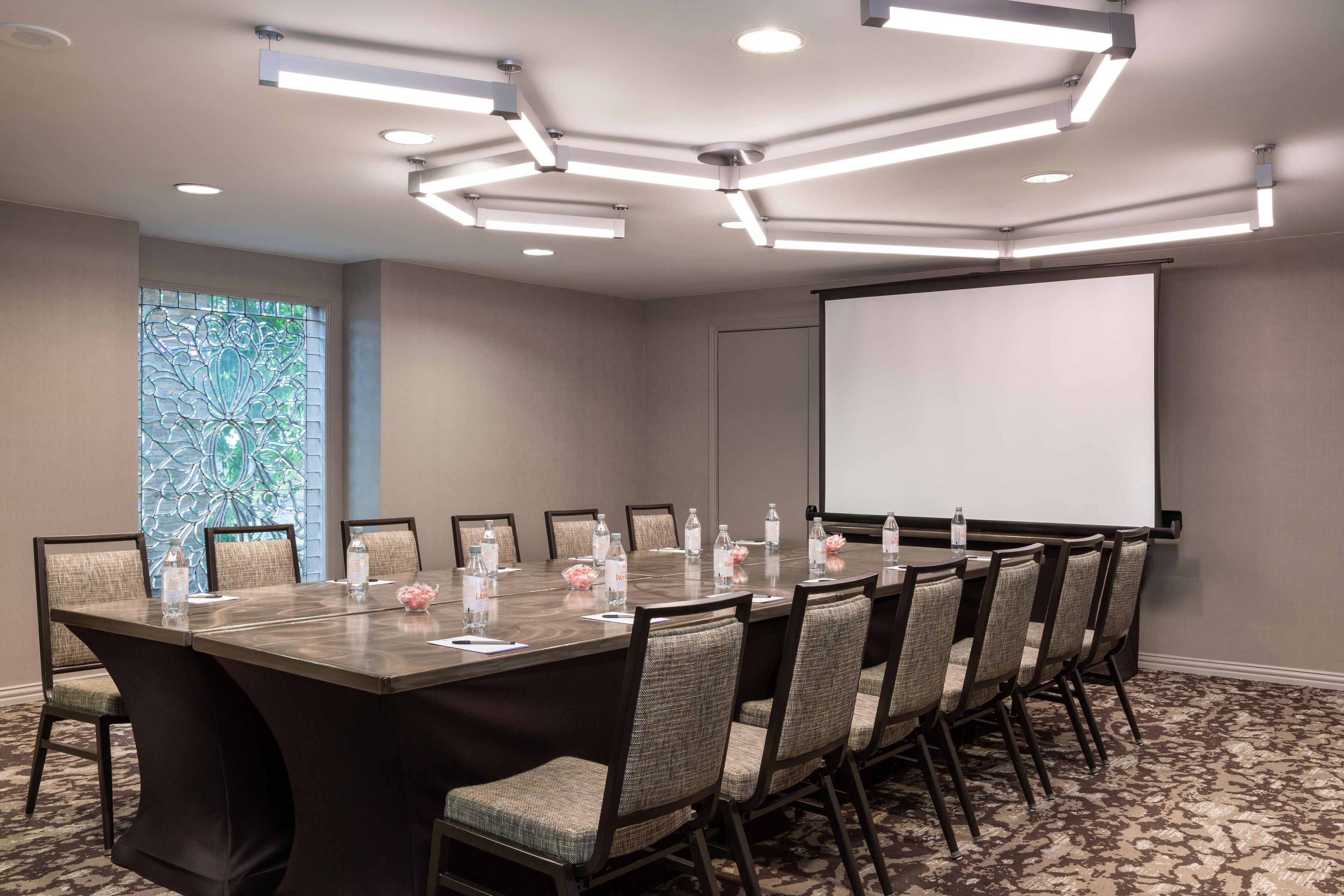 A conference room with a long table, chairs, a projector screen, and a decorative window.