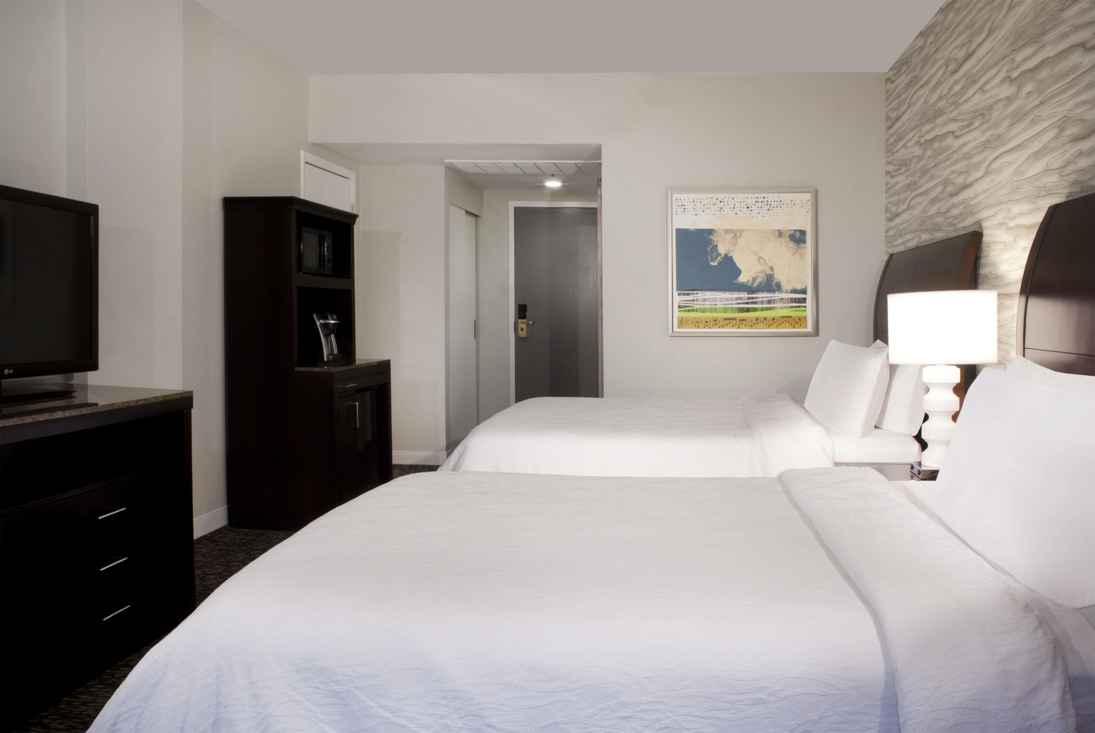 A hotel room with two beds, a television, a minibar, and a painting on the wall.