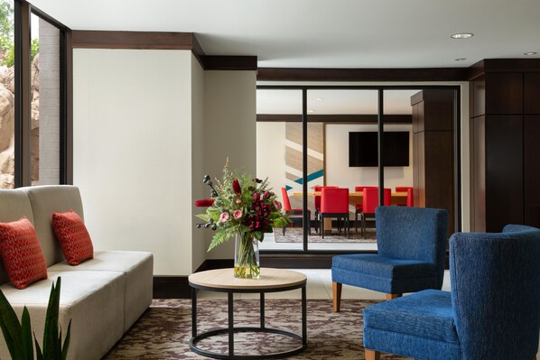Un moderno hall de hotel con un sofá, sillones azules, una mesa redonda con un florero, y vista a un comedor con sillas rojas.