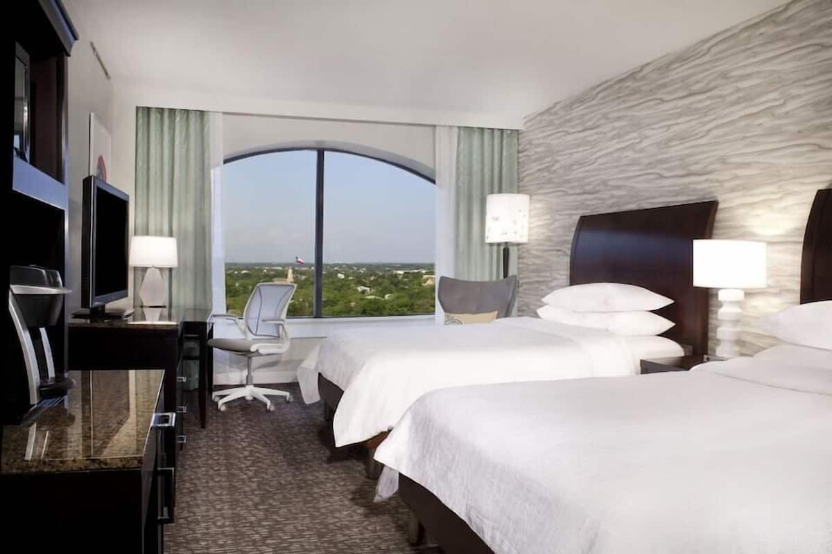 A hotel room with two beds, a desk, a chair, a TV, and a large window with a view.