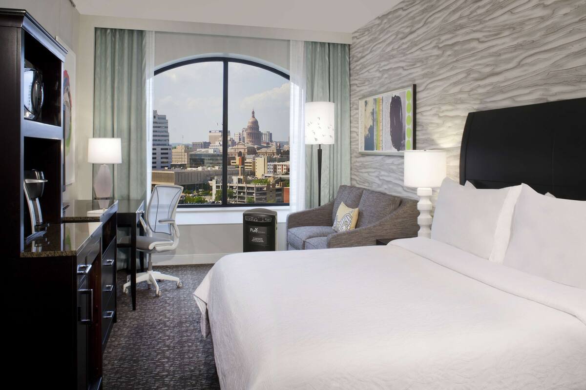 A hotel room with a large bed, a desk with a chair, a sofa, and a view of a cityscape through a large window.