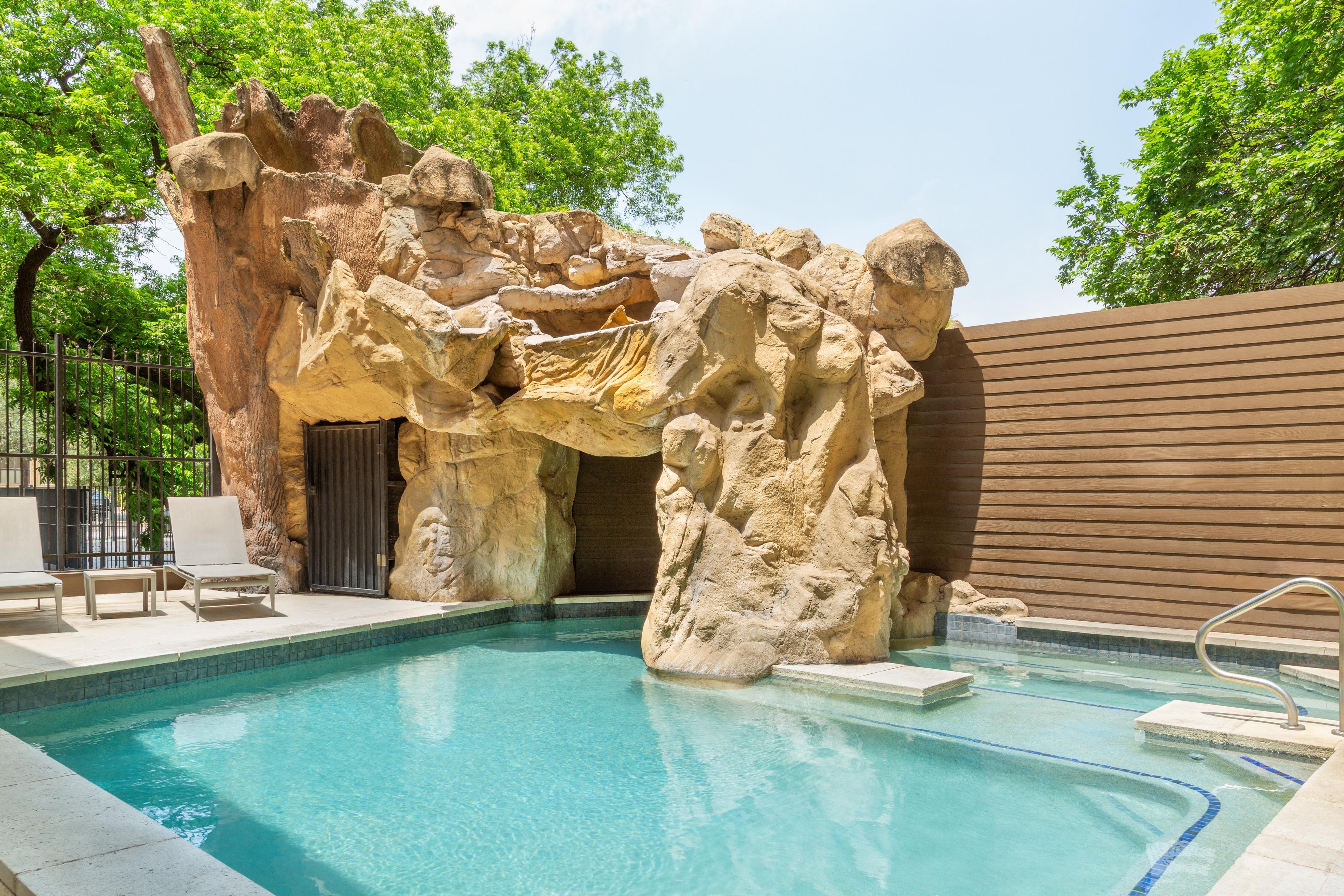 A pool with a natural rock formation, lounge chairs, and a wooden fence.