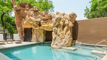 A pool with a natural rock formation, lounge chairs, and a wooden fence.