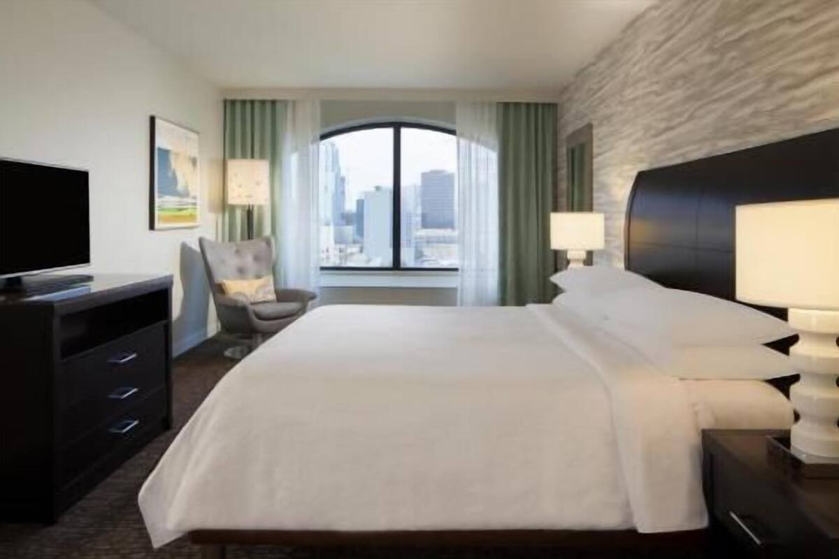 A hotel room with a bed, a TV, a desk, a chair, and a window with a view of buildings.