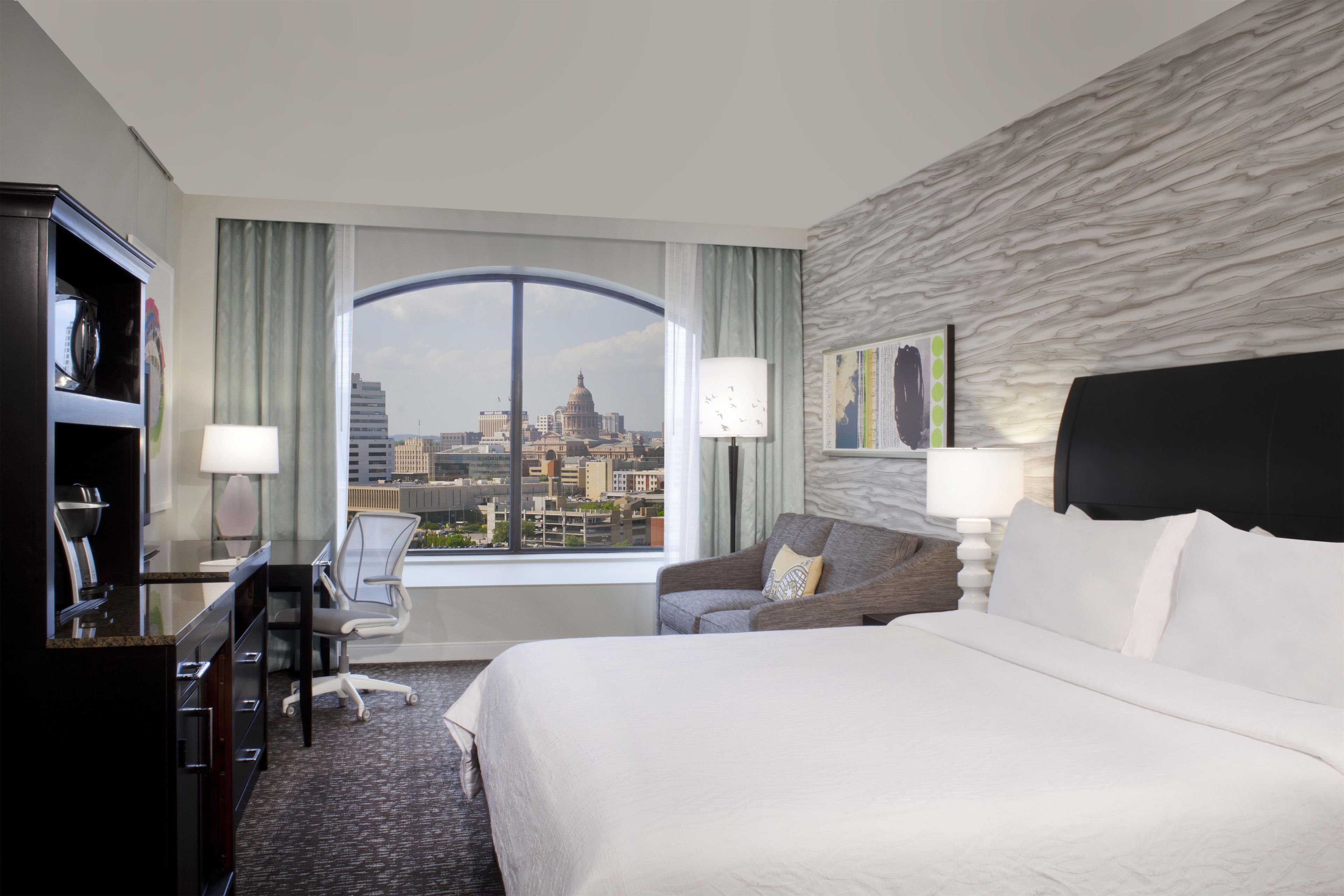 Een hotelkamer met een bed, een bureau, een stoel en uitzicht op een stadsgezicht door een groot raam.
