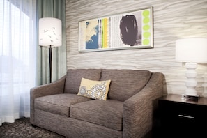 A modern living room with a grey sofa, a decorative pillow, a framed abstract artwork, a floor lamp, and a dark wooden side table. - Hilton Garden Inn Austin Downtown (Austin)
