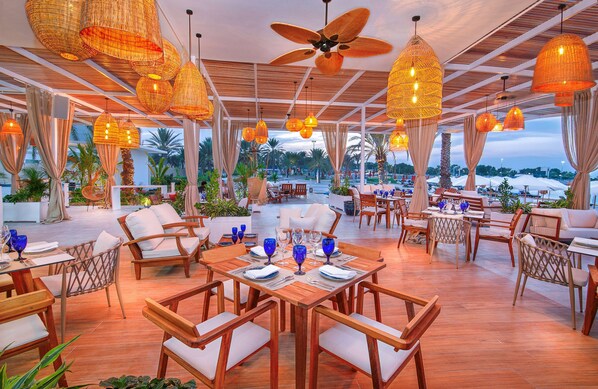 A restaurant with wooden tables and chairs, hanging wicker lamps, and a view of palm trees. - Radisson Blu Hotel & Resort, Abu Dhabi Corniche (Abu Dhabi)