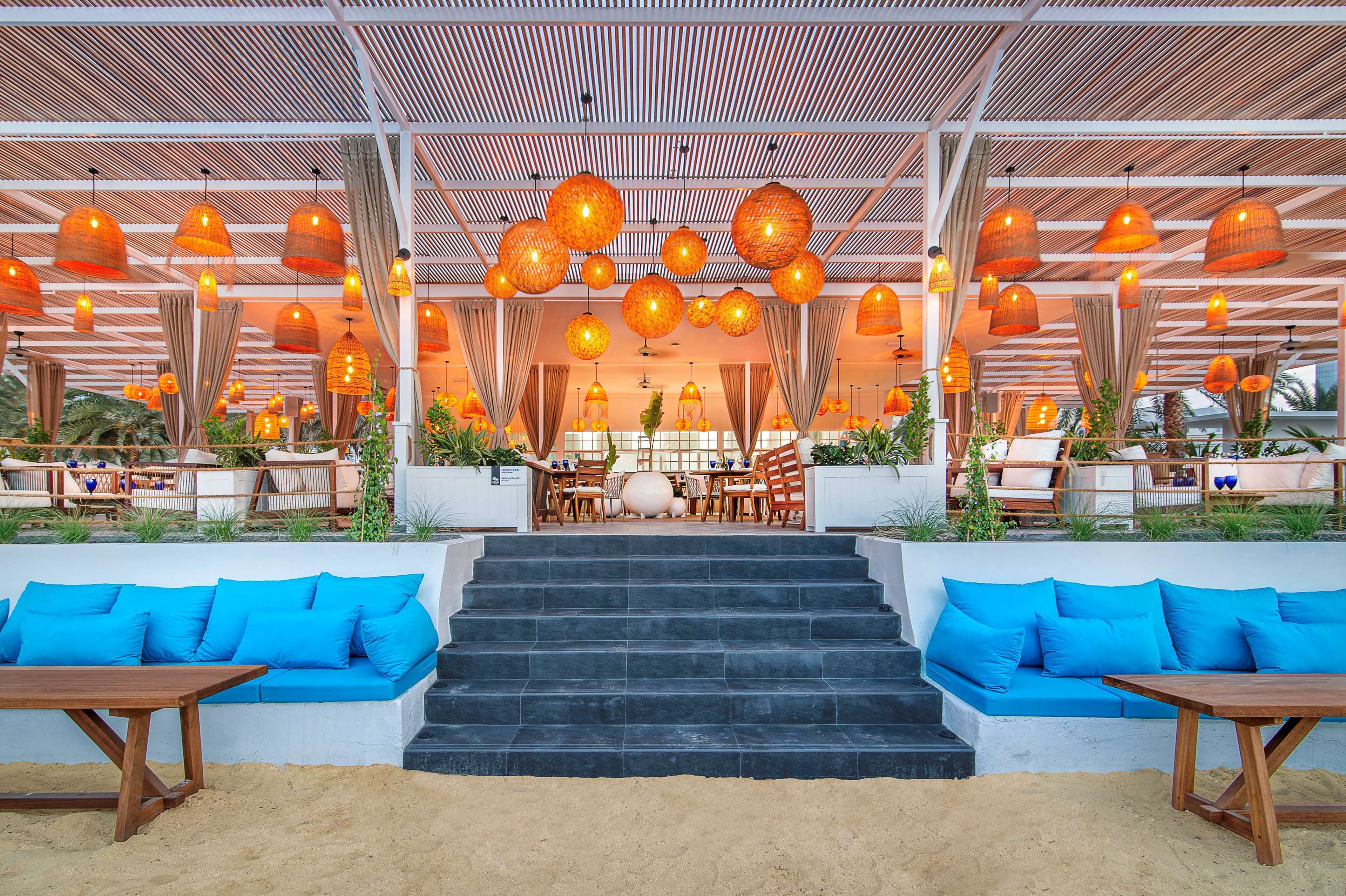 A covered outdoor seating area with blue sofas, a staircase leading to an upper level, and hanging orange pendant lights.