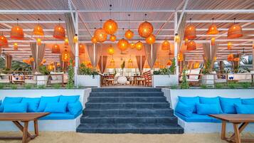 Ein überdachter Außenbereich mit blauen Sofas, einer Treppe zu einer erhöhten Ebene und hängenden, orangefarbenen Pendelleuchten.
