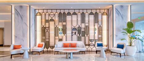 Un lobby moderno con piso de mármol, una zona de estar central con un sofá y sillones, y una pared decorativa con patrones geométricos.