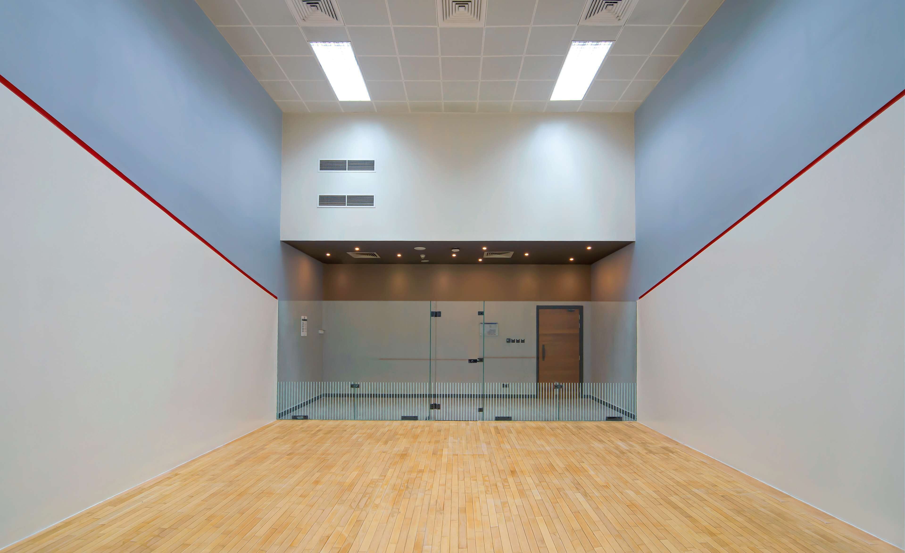 A spacious indoor squash court with a wooden floor, white walls, and a red line marking the playing area.