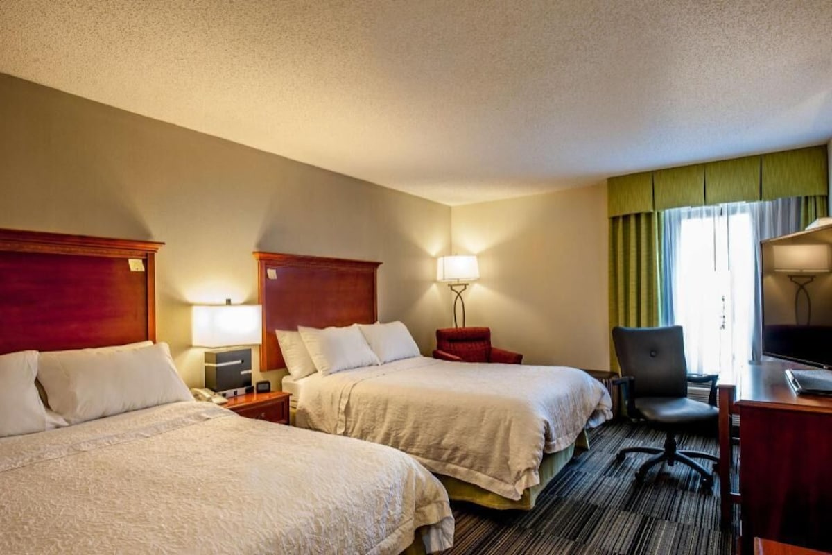 A hotel room with two beds, a desk, a chair, and a television.