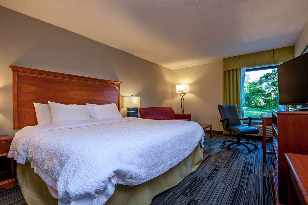 Ett hotellrum med en stor säng, ett skrivbord med en stol, en TV och ett fönster med utsikt över grönområden.