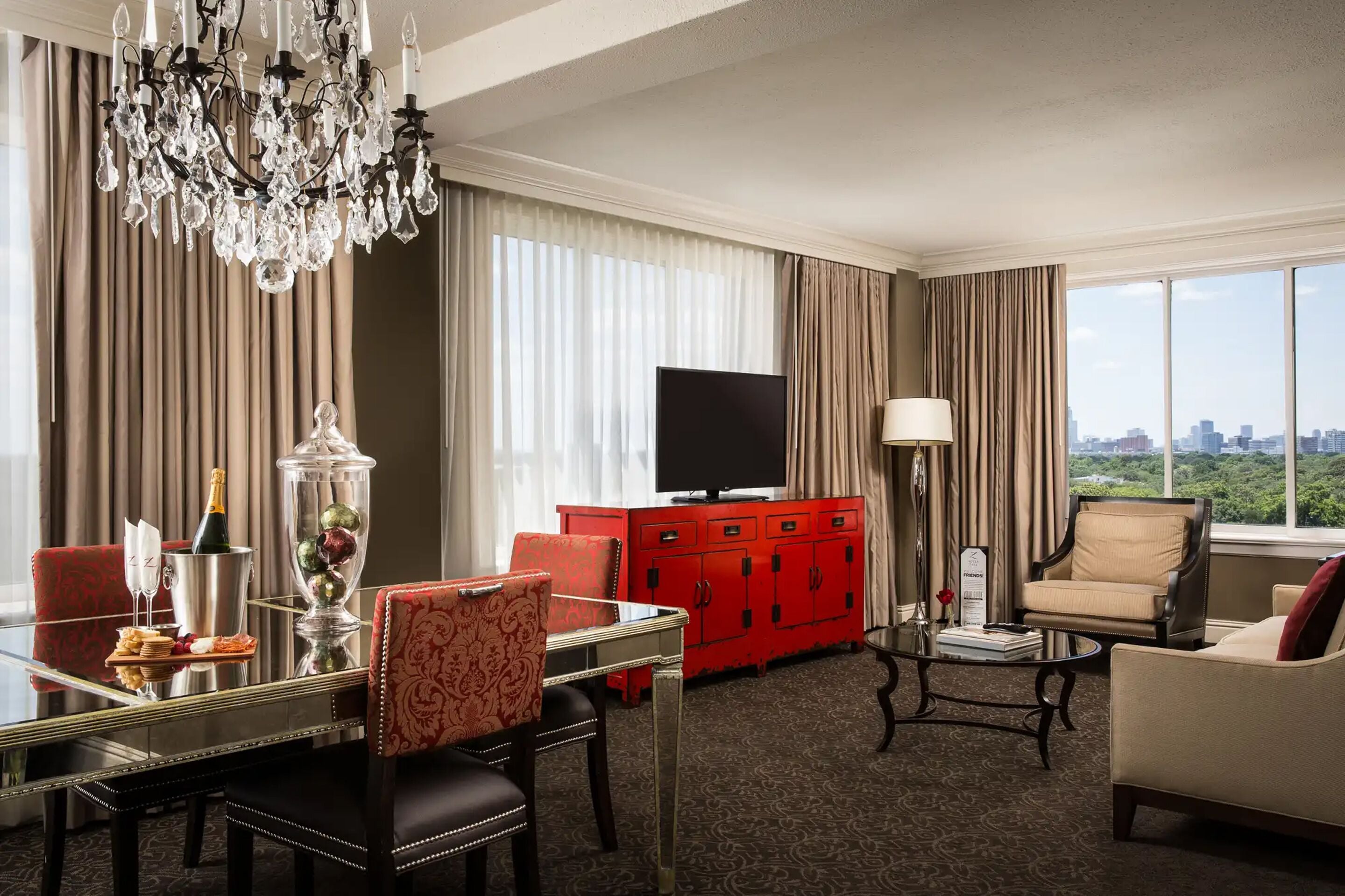 A hotel room with a large window, a chandelier, a red cabinet, a television, and a dining area.